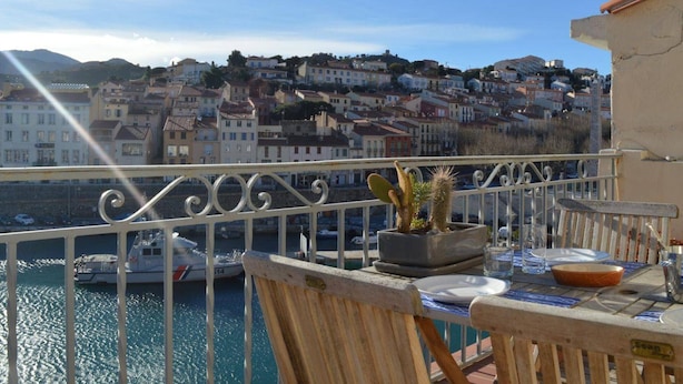 A balcony with a table set for dining, overlooking a marina and a hillside town.