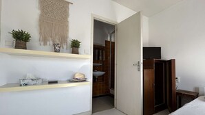 A bedroom with a bed, a wooden wardrobe, a shelf with toiletries, and a macrame wall hanging.