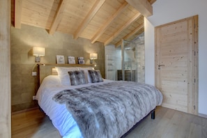 Quarto com teto de madeira, cama com manta de pele, mesa de cabeceira com luminária e banheiro visível através de uma porta de vidro.