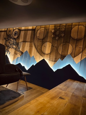 Quarto com painel de parede de madeira retratando montanhas e corpos celestes, uma cadeira moderna e piso de madeira.