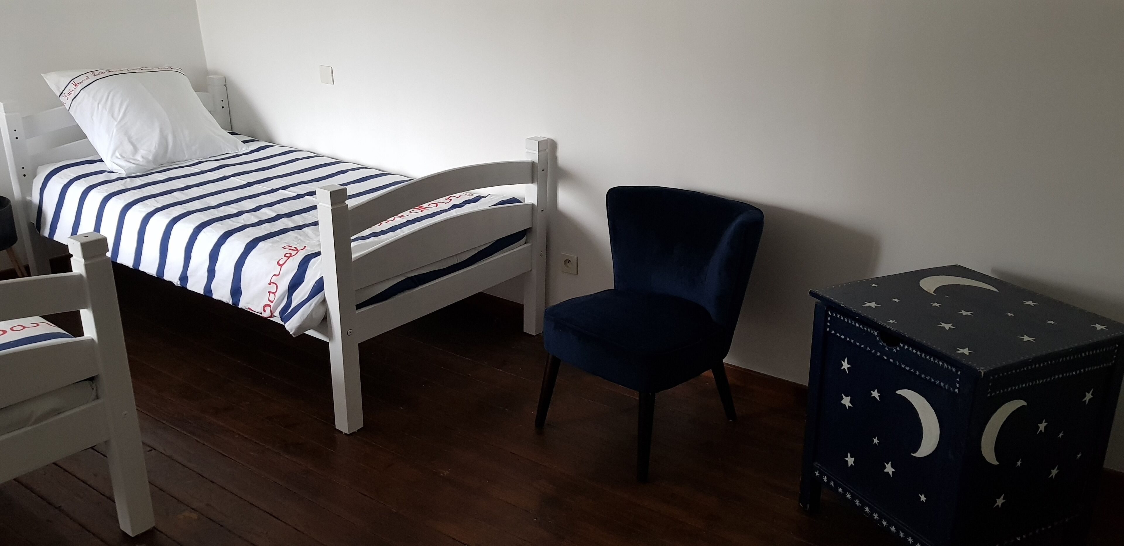 A single bed with a striped bedspread, a dark blue chair, and a small table with a star and moon design.