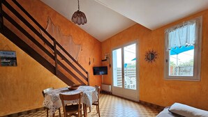 Huoneessa on kalanruotokuviollinen lattia, ruokapöytä tuoleineen, sänky, televisio ja ikkuna, jossa on verhot.