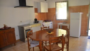 Eine kleine Küche mit Herd, Backofen und Kühlschrank. Ein Esstisch mit Stühlen und ein Fenster mit Vorhängen sind ebenfalls vorhanden.
