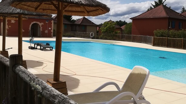 En swimmingpool med solstolar och ett parasoll med halmtak, omgiven av trästaket och byggnader.