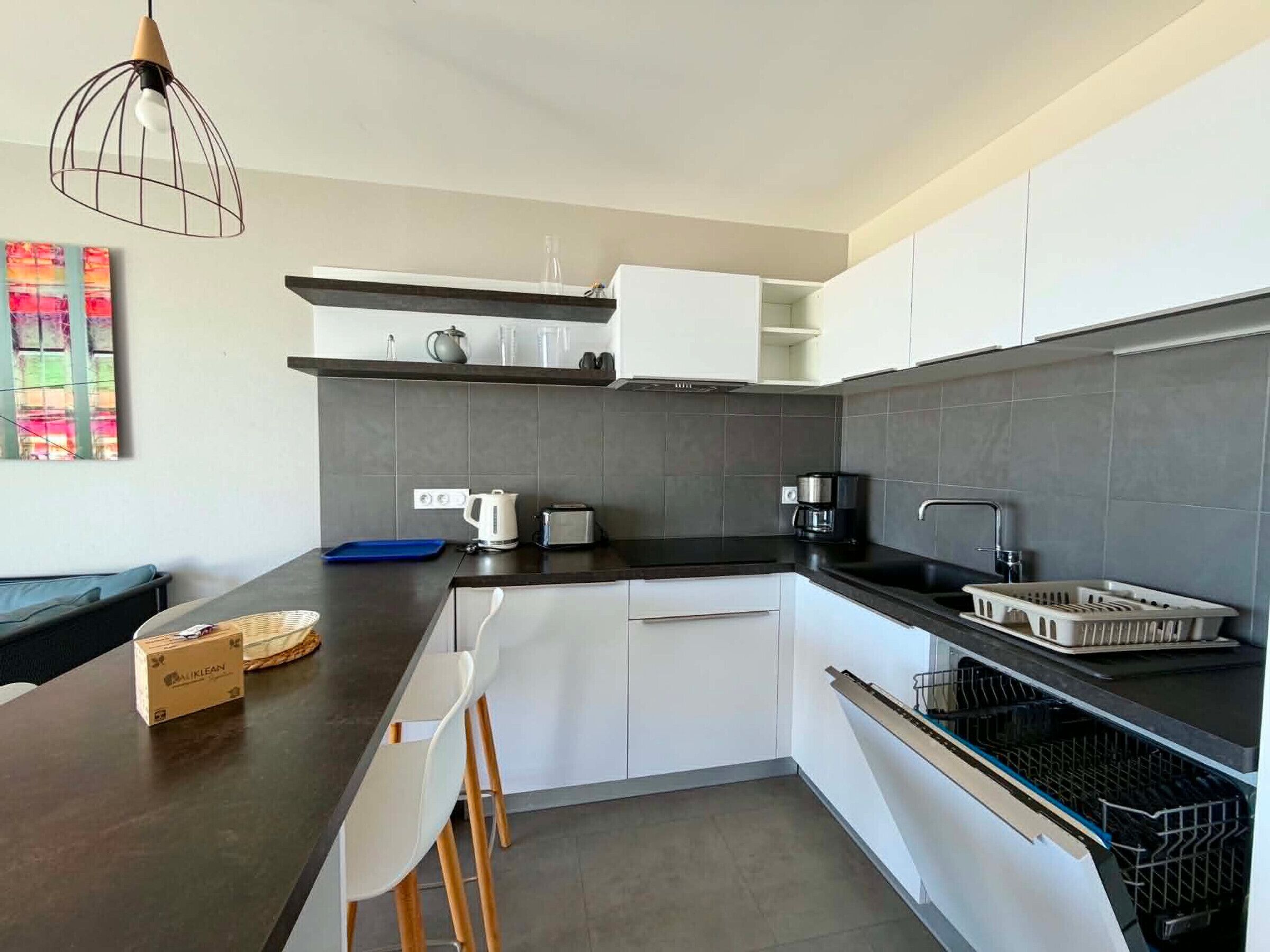 A modern kitchen with white cabinets, a black countertop, a coffee maker, a toaster, and a hanging light fixture.