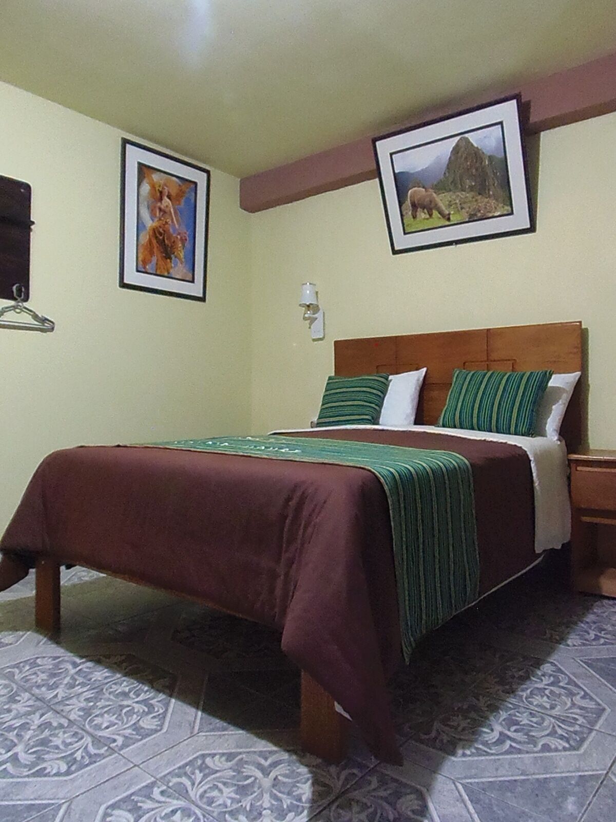 A bedroom with a wooden bed, green and white bedding, patterned floor tiles, and two framed pictures on the walls.