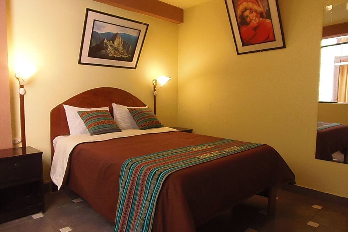 Quarto de hotel com cama, duas luminárias de apoio, uma fotografia emoldurada de uma montanha e um retrato pendurado na parede.