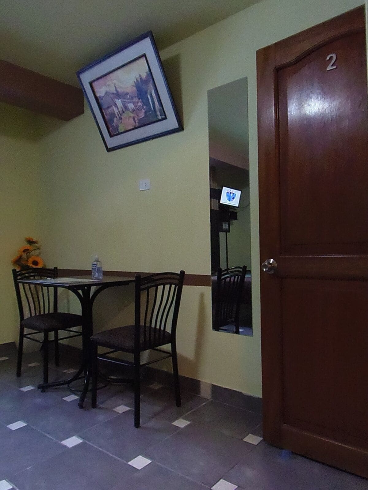 Un comedor con una mesa y sillas, un cuadro enmarcado en la pared, una puerta con el número 2 y un piso de baldosas.