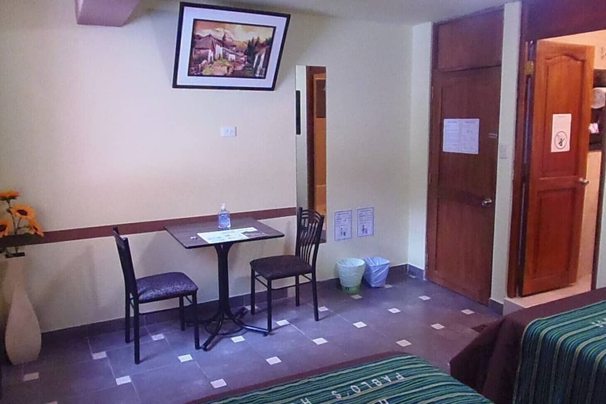 Quarto de hotel pequeno com uma cama de solteiro, uma mesa com duas cadeiras, um quadro na parede e uma lixeira.