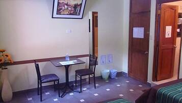 Une petite chambre d’hôtel avec un lit simple, une table avec deux chaises, un tableau encadré au mur et une poubelle.