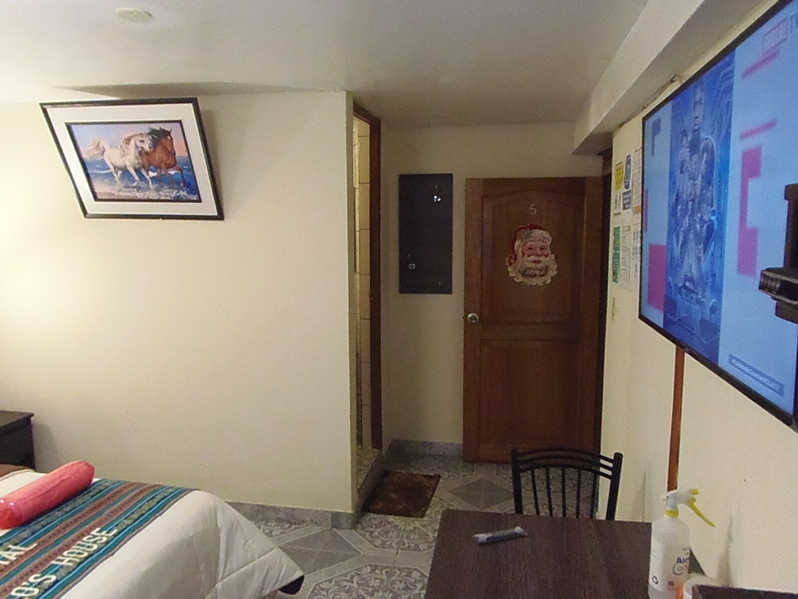 A hotel room with a bed, a small table, a chair, a picture on the wall, and a door with a Santa Claus decoration.