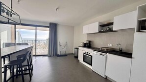 A modern kitchen with white cabinets, a black countertop, and a view of the sea through a large window. - Luxury apartment for 4, beachfront, balcony, pool, pets allowed, parking, WiFi (La Tranche-sur-Mer)