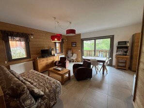 A cozy cabin-style room with a sofa, a coffee table, a TV, a dining table with chairs, and a large window offering a view of greenery. - Spacious 3* Apartment - 6 ppl with balcony in Châtel (Châtel)