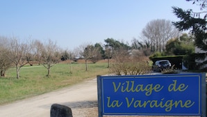 Una señal que dice 'Village de la Varaigne' en una carretera rural con árboles y un coche estacionado al fondo.