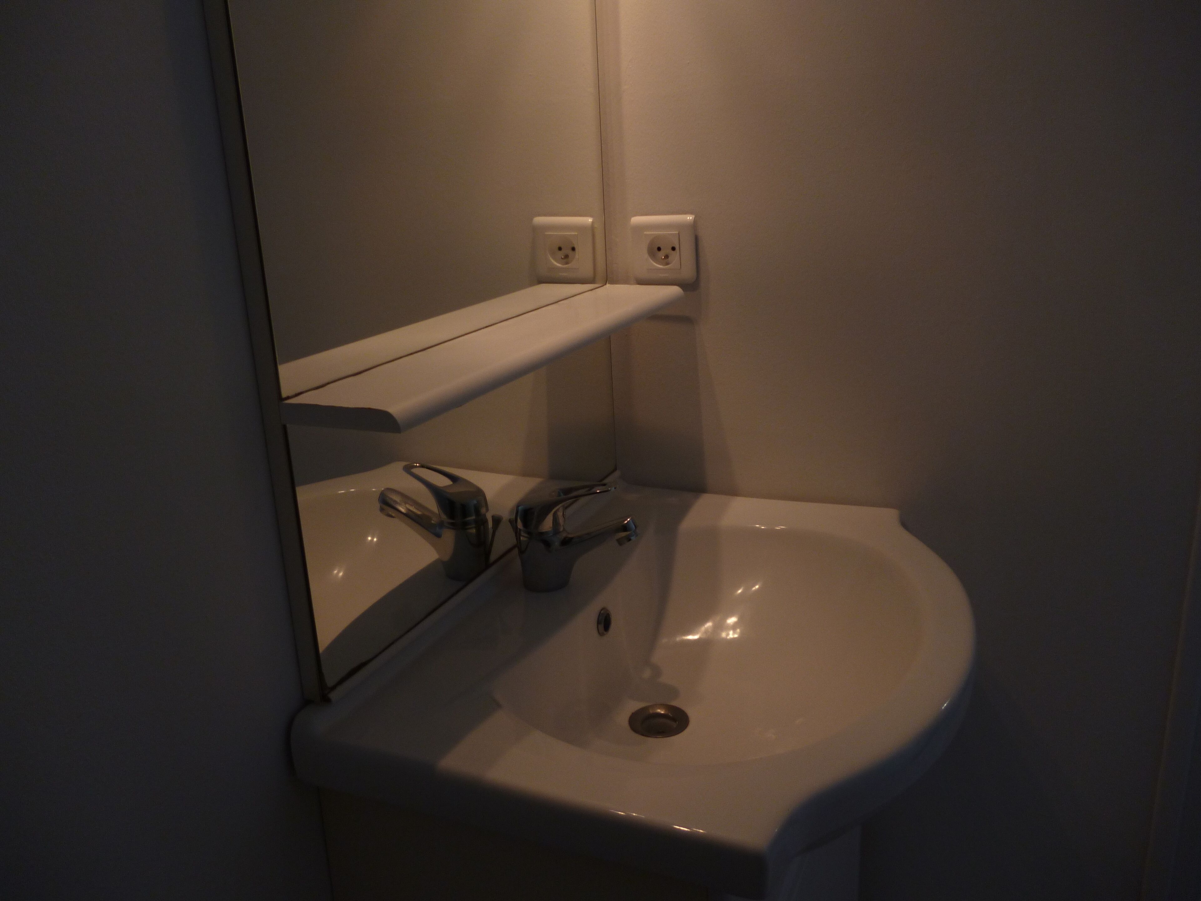 Une salle de bain avec un lavabo blanc, un miroir et deux prises électriques au mur.