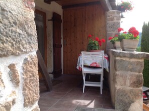 A stone building with a wooden door, a white chair, a table with a red tablecloth, and potted red flowers.