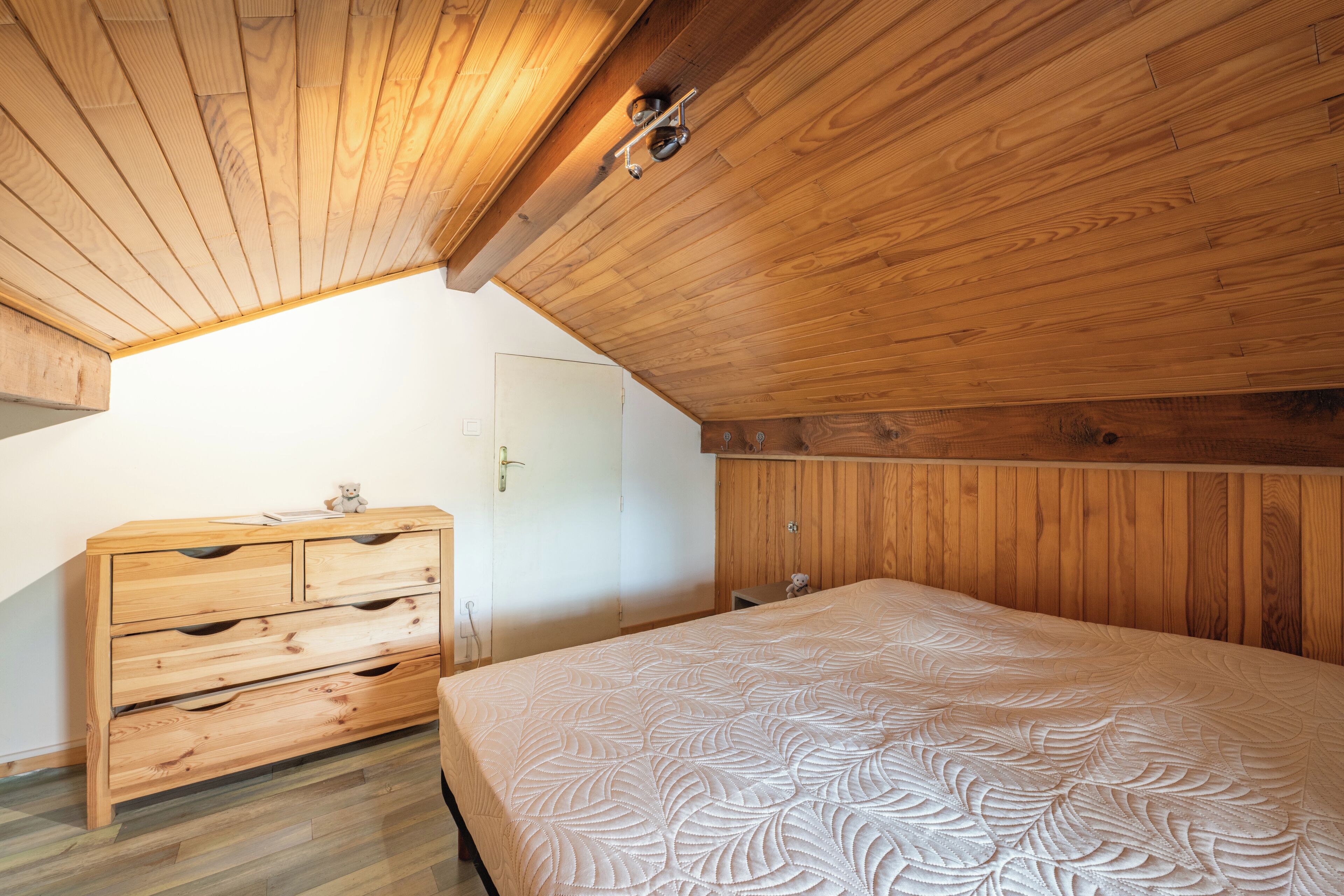 Ein Schlafzimmer mit schrägen Wänden, Holzwänden, einem Bett mit gemustertem Überwurf, einem Holzschrank und einer Tür.