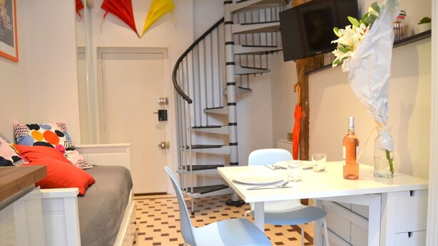 Un espacio habitable pequeño con zona de comedor, un sofá con cojines de colores, una escalera y un televisor montado en la pared.