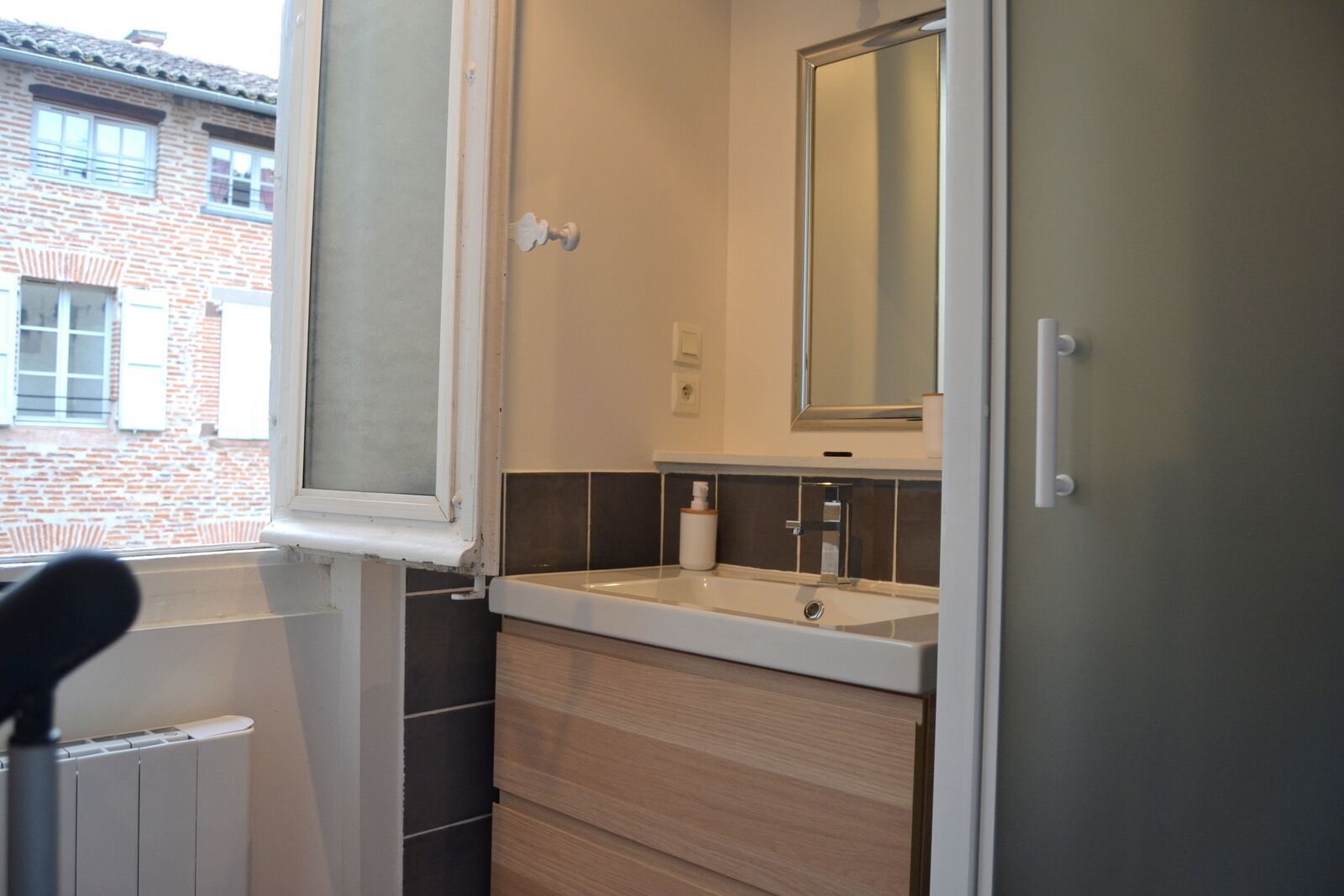 Een badkamer met een wit wastafelmeubel, donkere tegels, een spiegel en een raam met uitzicht op een bakstenen gebouw.