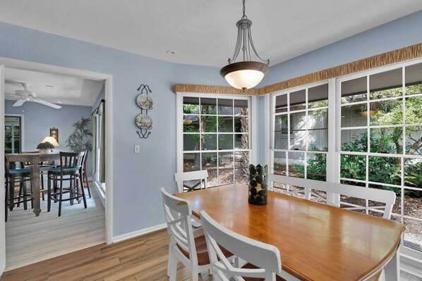 A dining area with a wooden table and white chairs, large windows with a view of greenery, and a ceiling fan.