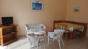Una habitación con una televisión sobre un soporte de madera, una silla de mimbre con cojín, una mesa de comedor con sillas y un cuadro enmarcado en la pared.
