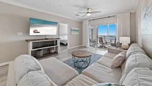 A spacious living room with a large flat-screen TV, a ceiling fan, and a view of the ocean through large windows. - Come Enjoy this luxury condo right on the beach of Gulf Shores! (Orange Beach)