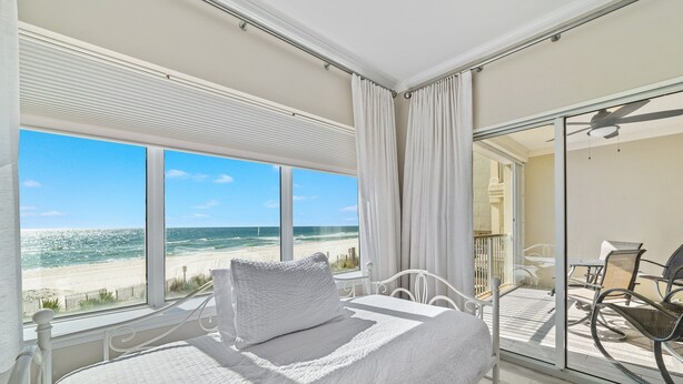 A bedroom with a large window overlooking a beach, a balcony with outdoor furniture, and a ceiling fan. - Come Enjoy this luxury condo right on the beach of Gulf Shores! (Orange Beach)