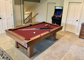 Una mesa de billar con bolas y tacos, una escalera y una consola de videojuegos en una habitación con suelo alfombrado y puertas blancas.