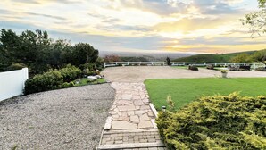 Un camino pavimentado que lleva a una cerca blanca con vista a un césped verde y un paisaje urbano distante.