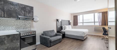 A modern hotel room with a kitchenette, a bed, a sofa, and a large window.