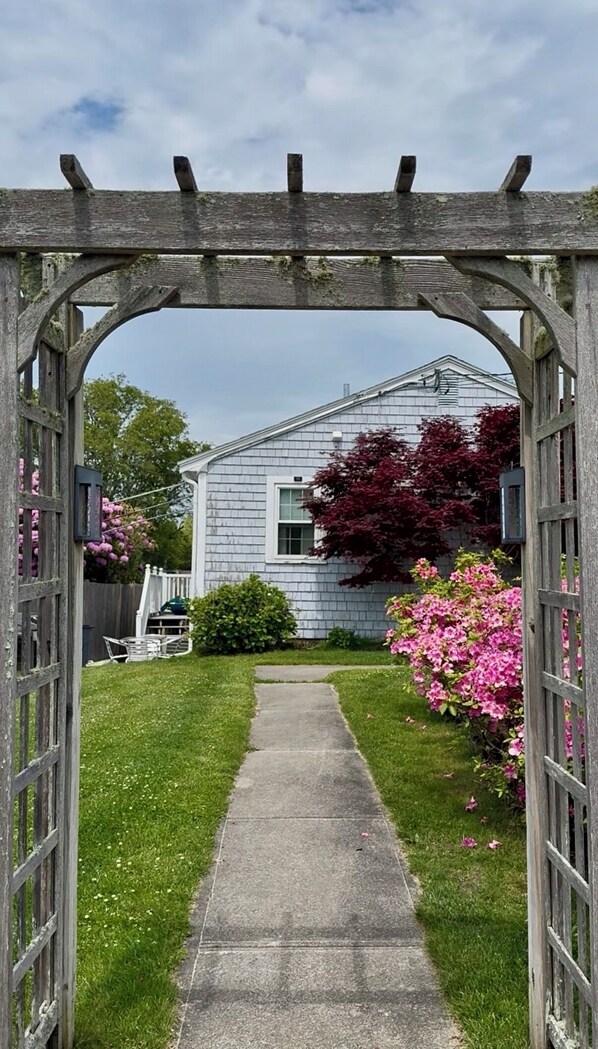 A wooden arbor leading to a house with a white exterior and a red-leafed tree.