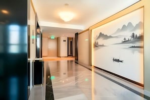 Een hotelgang met een spiegelende vloer, een groot wanddecor dat een vredig landschap toont en een moderne lamp aan het plafond.