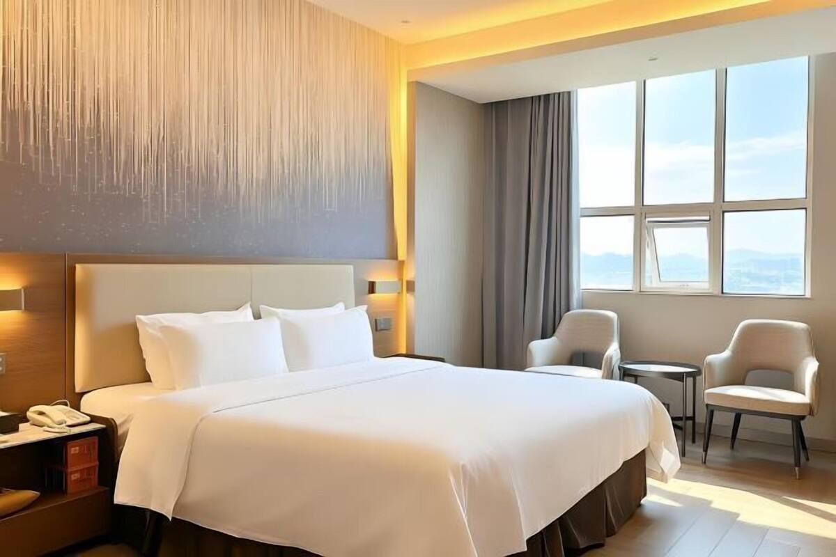 A hotel room with a large bed, two chairs, a small table, a nightstand, and a view of the outdoors through large windows.