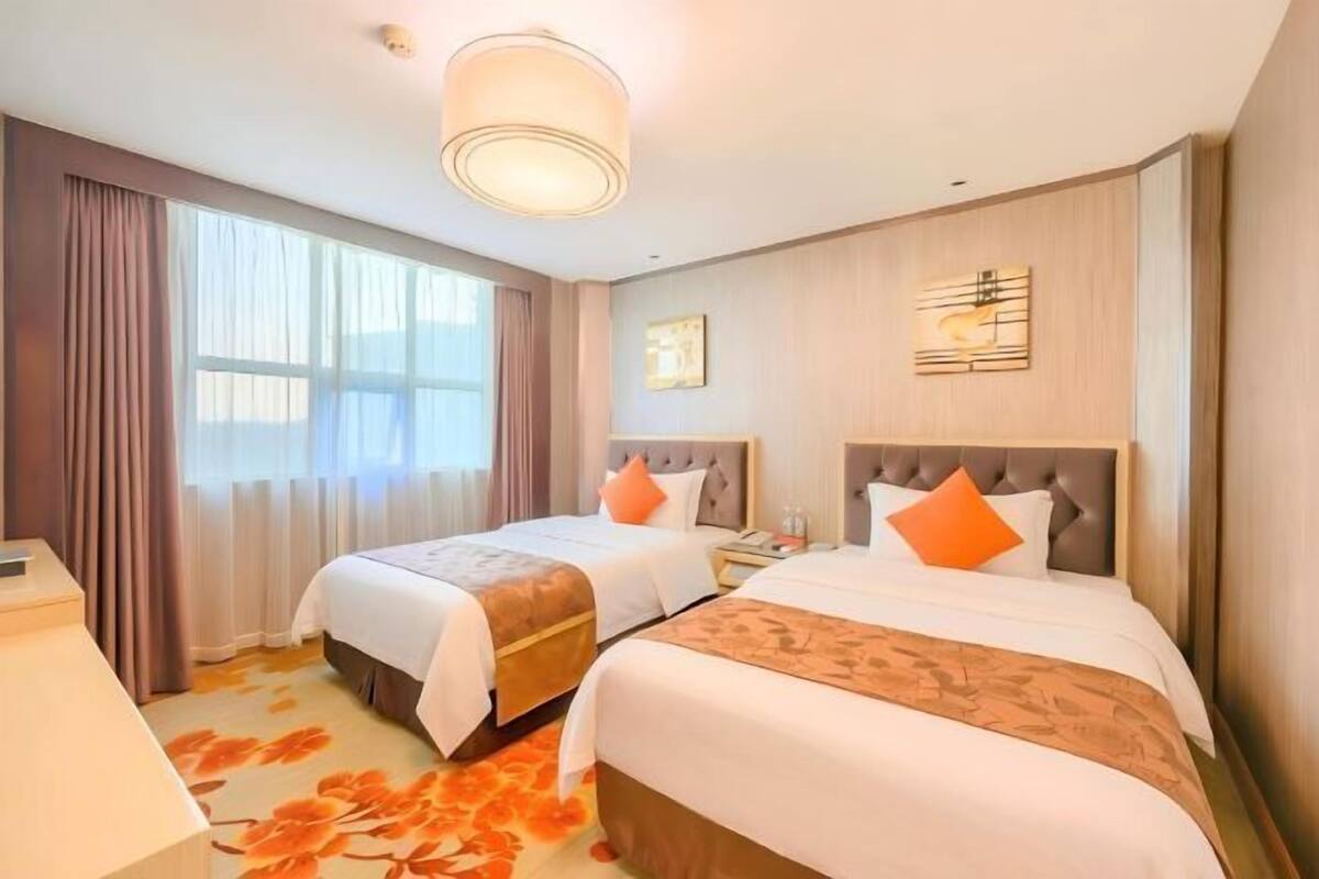 A hotel room with two beds, a large window with curtains, a wall-mounted artwork, and a carpet with a floral pattern.