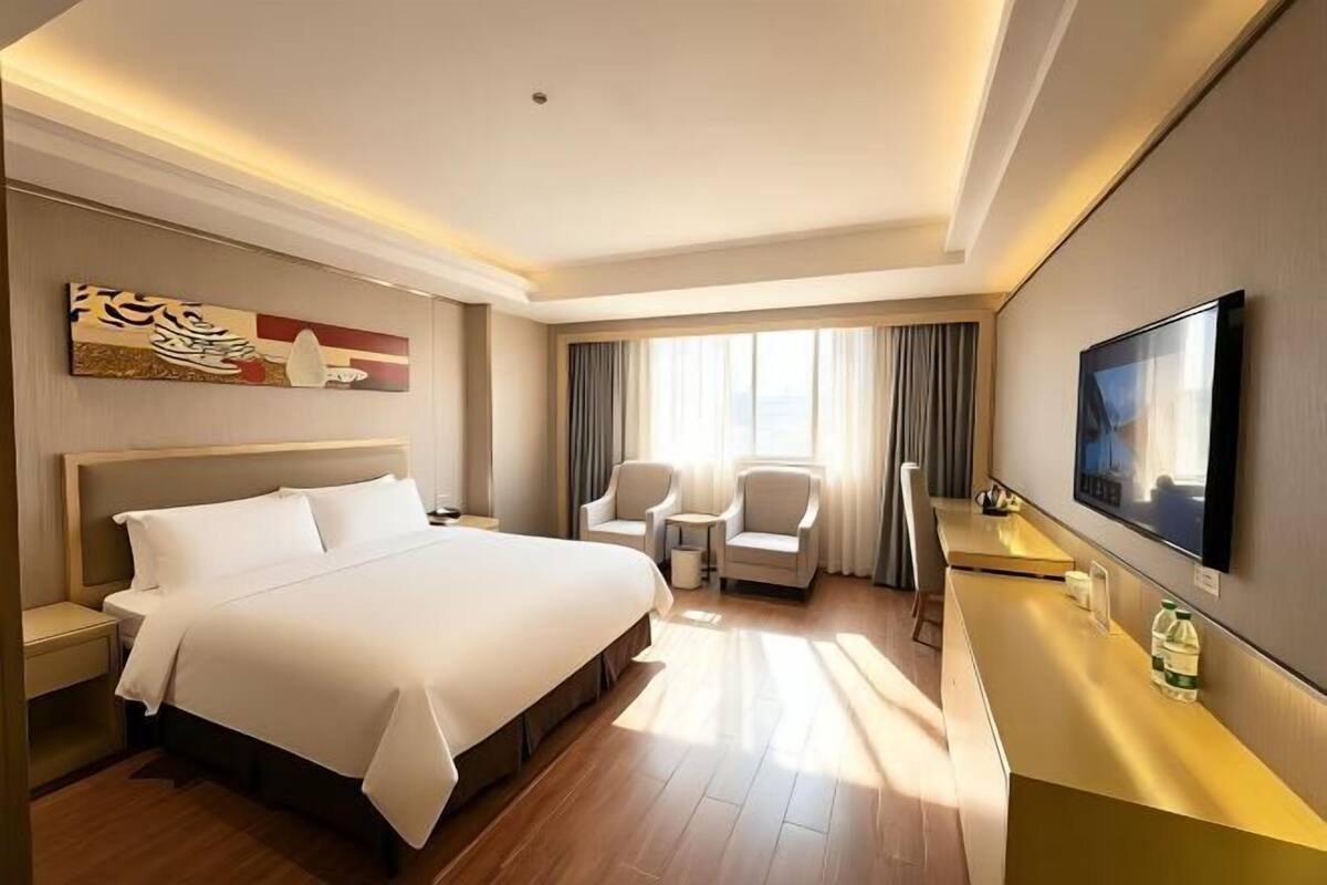 A hotel room with a large bed, two chairs, a desk, a flat-screen TV, and a large window with curtains.