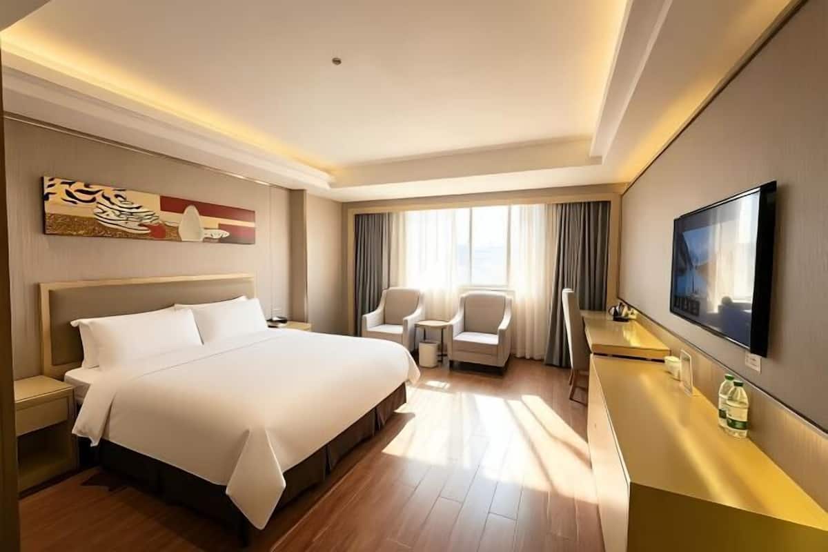 Quarto de hotel com uma cama grande, duas cadeiras, uma escrivaninha, televisão de tela plana e uma janela ampla com cortinas.