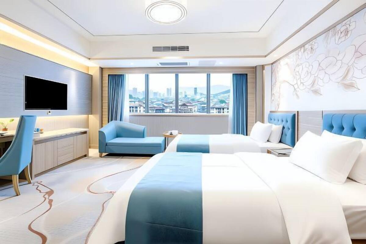 A hotel room with two beds, a desk, a chair, a TV, and a large window with a city view.
