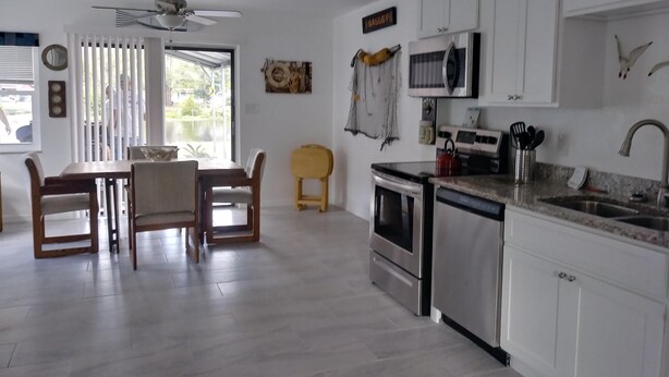A modern kitchen with white cabinets, a dining area with wooden chairs, a ceiling fan, and a view through the window. - WATERFRONT COTTAGE w/ACCESS TO WEEKI WACHEE RIVER & GULF FRESHLY REMODELED! (Weeki Wachee)