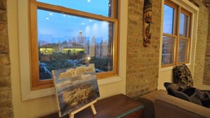 Una habitación con un ventanal grande que da a un paisaje urbano, una mesa pequeña con un libro titulado "About Chicago" y una pared de ladrillo con un adorno.