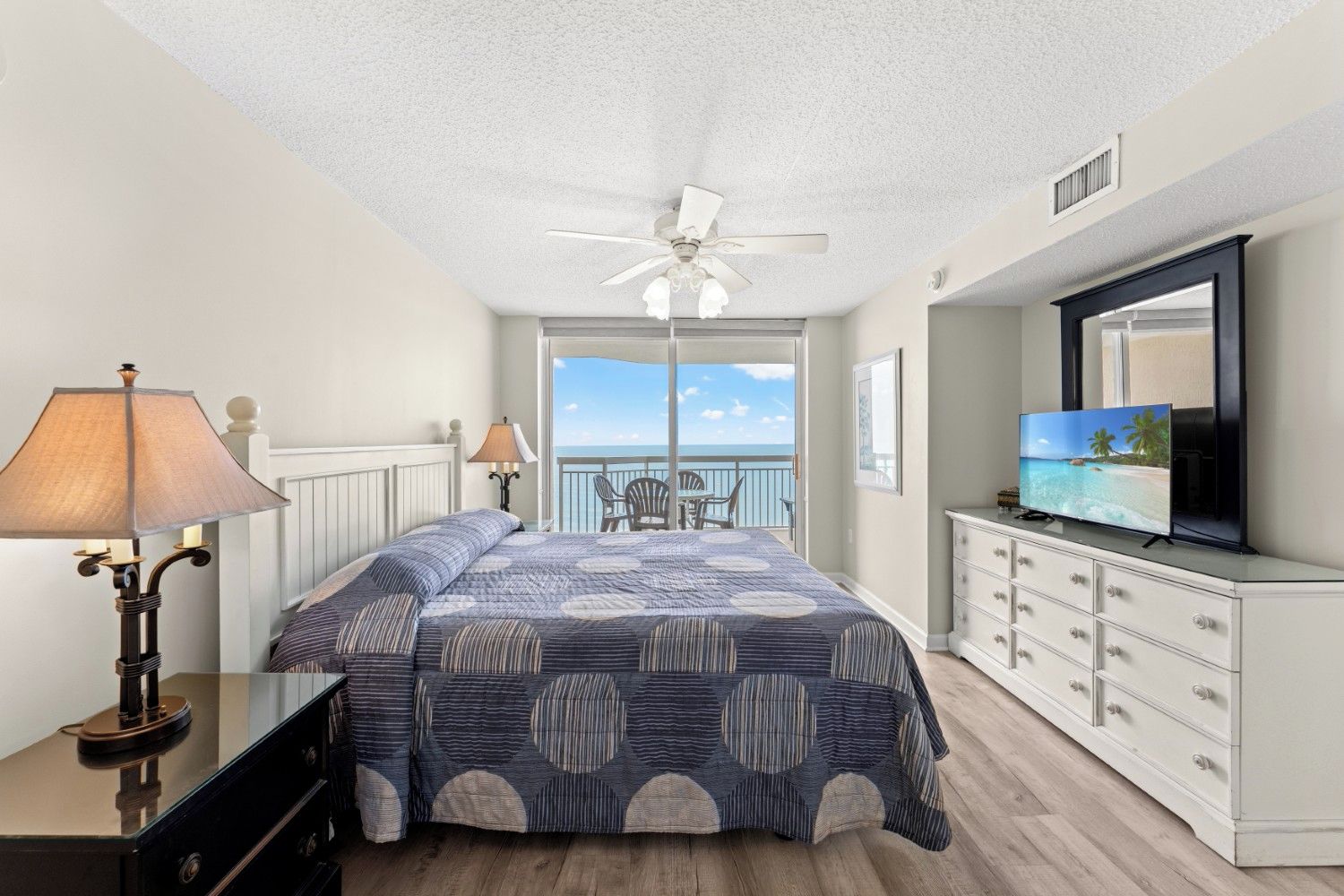 A bedroom with a bed, a ceiling fan, a TV, and a view of the ocean.