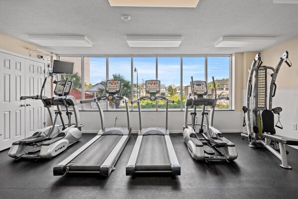 Un gimnasio con cintas de correr, elípticas y máquinas de pesas, ventanas amplias con vistas a casas, y una puerta con cámara de seguridad.