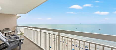 A balcony with a view of the beach, a table with chairs, and a railing.