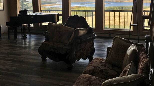 Un'ampia stanza con grandi finestre che offrono vista su un lago e montagne lontane. Si trova un pianoforte, una poltrona con motivi decorativi e una lampada da terra.