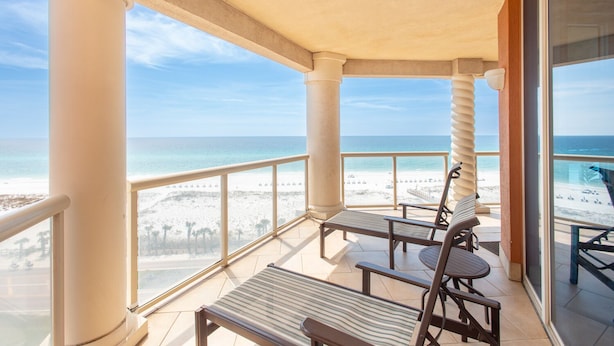 A balcony with a view of the beach, two lounge chairs, and a table.