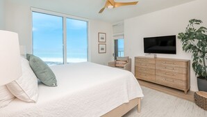A bedroom with a large bed, a ceiling fan, a TV, a dresser, and a plant.