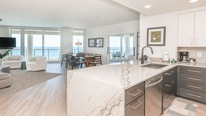 A modern kitchen with a marble island, dark cabinetry, and a large window overlooking the ocean.