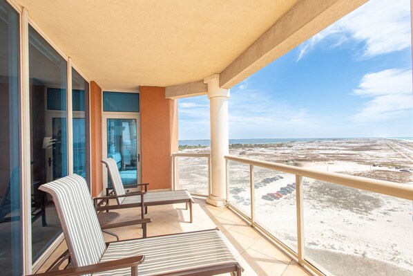 A balcony with two striped lounge chairs, a glass door, and a view of a beach with parked cars.