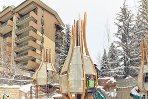 Un télésiège avec une personne assise, entouré d’arbres couverts de neige et d’un immeuble à plusieurs étages doté de balcons.