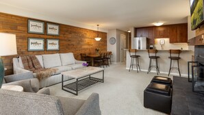 Un salon moderne comprenant un canapé, une table basse et un pouf, un coin repas avec une table et des chaises, et une cuisine avec des meubles en bois et un réfrigérateur.