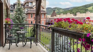 Un balcon avec deux chaises et une table, donnant sur un jardin aux fleurs multicolores et offrant une vue sur des bâtiments et des collines.